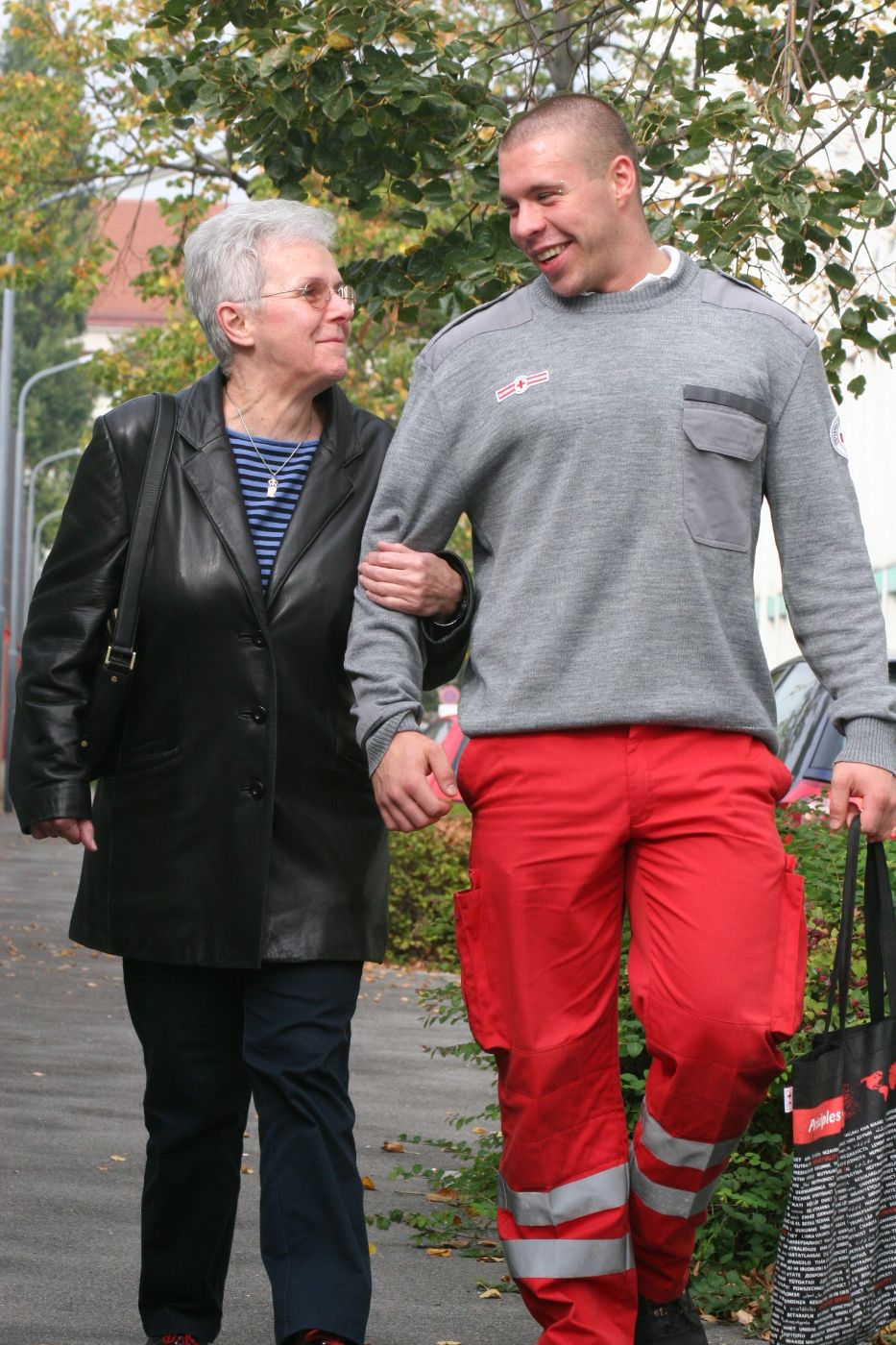 Ein Zivildiener in einer Uniform des Österreichischen Roten Kreuzes begleitet eine ältere Dame bei einem Spaziergang. Der Zivildiener trägt dabei eine Tasche.