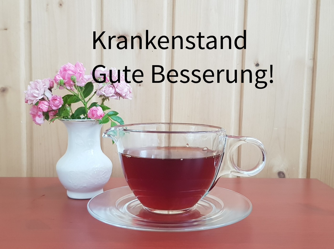 Symbolfoto mit einer Tasse aus Glas mit Tee, links daneben steht eine weiße Vase mit rosaroten Blumen. Die Tasse Tee steht auf einem roten Tisch.